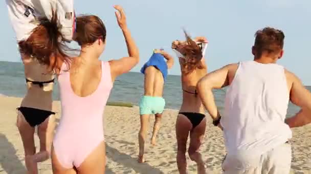 Visão traseira do grupo de amigos despir correndo para a água do mar no pôr do sol e jogar camisas na praia. Pessoas felizes alegres se divertindo na praia. Homens e mulheres vão nadar em câmera lenta . — Vídeo de Stock