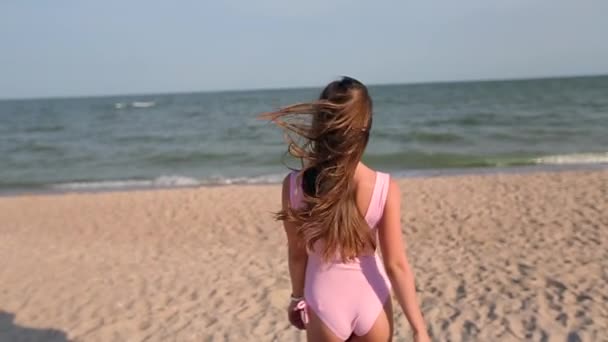 Back view of woman walking on beach to the sea water, turns and invites to follow her on vacation travel. Lady with slim hot body on tropical white sand beach wearing pink swimsuit goes to the ocean.