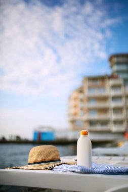 Plaj havlusu güneş krem şişesinde deniz kıyısında arka plan ile. Lüks spa Resort gündoğumu veya günbatımı üzerinde açık havada şezlong üzerinde güneş kremi. Cilt bakım ve koruma kavramı ve seyahat. Altın bronzluk.