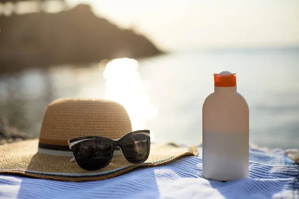 Suntan cream bottle and sunglasses on beach towel with sea shore on background. Sunscreen on deck chair outdoors on sunrise or sunset. Skin care and protection concept. Golden tan. — Stock Photo, Image