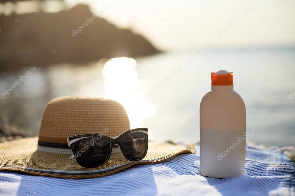 Suntan cream bottle and sunglasses on beach towel with sea shore on background. Sunscreen on deck chair outdoors on sunrise or sunset. Skin care and protection concept. Golden tan.