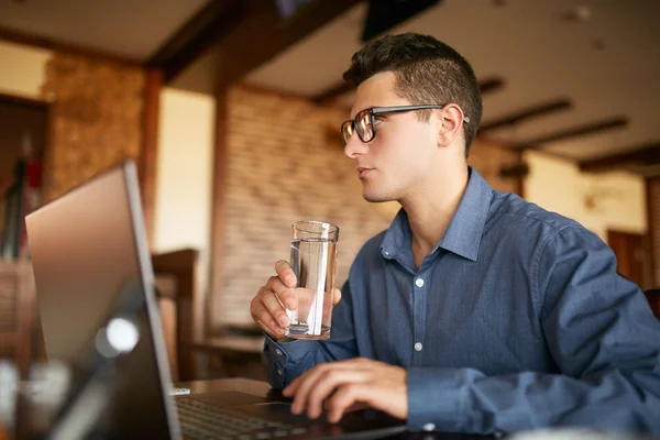 Handsome caucasian man with glass of water in hand works with laptop. Businessman in glasses drinks water for body hydration while working. Attractive designer quench thirst. Healthy lifestyle theme. — Stock Photo, Image