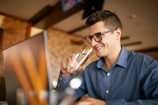 Smiling caucasian man with glass of water in hand works with laptop. Businessman in glasses drinks water for body hydration while working. Attractive designer quench thirst. Healthy lifestyle theme. — Stock Photo, Image