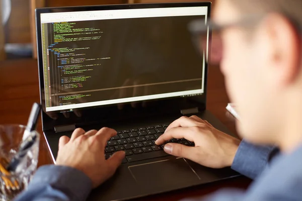 Back view over the shoulder shot of developer programmer with laptop. Program code and script data on the screen. Young freelancer in glasses working on project in cafe.