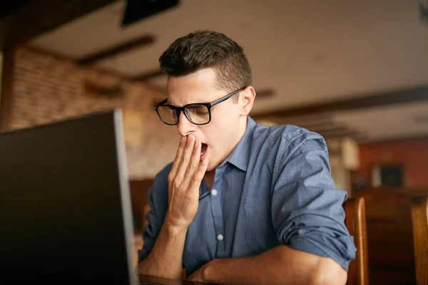 Early morning in the office. Sleepy tired handsome hipster freelancer in glasses is yawning at his work place in front of the laptop screen on desk. Overworked businessman in office. — Stock Photo, Image