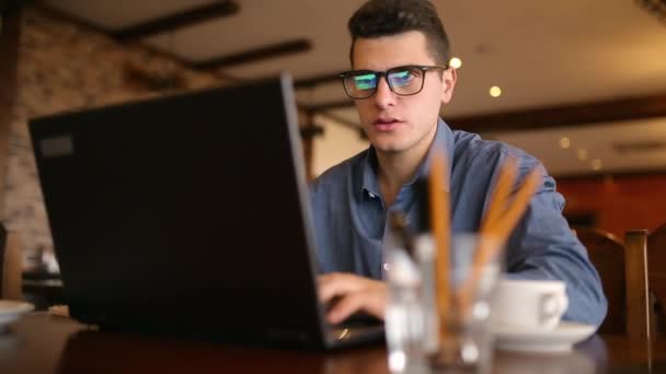 Όμορφος freelancer επιχειρηματίας σε ποτήρια εργάζονται επιμελώς για φορητό υπολογιστή στην καφετέρια και, στη συνέχεια, ποτά φλιτζάνι του καφέ. Ο άνθρωπος που εργάζονται με το pc σε coffeeshop. Επιχειρηματική ιδέα multitasking. Ρυθμιστικό πλάνο παρακολούθησης — Αρχείο Βίντεο