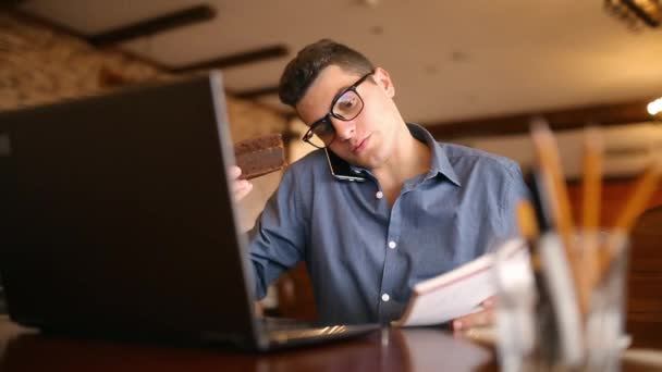 Ein gutaussehender junger Geschäftsmann mit Brille arbeitet am Laptop, telefoniert, macht Notizen und sucht Informationen in einem Notizbuch, trinkt Kaffee und isst einen Kuchen. Multitasking-Konzept für die Telekommunikation. — Stockvideo