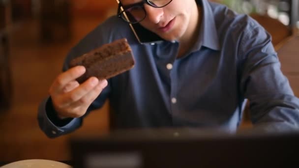 Harried handsome young businessman in glasses working on laptop, speaking on phone, taking notes and searches info in notebook, drinking coffee and eating a cake. Multitasking telecomuting concept. — Stock Video