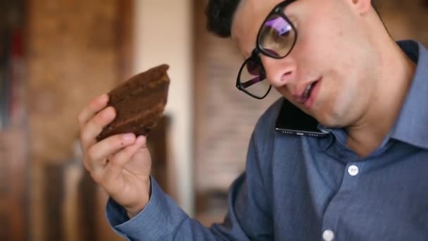 Harried joven hombre de negocios guapo en gafas que trabajan en el ordenador portátil, hablando por teléfono, tomando notas y busca información en el cuaderno, beber café y comer un pastel. Concepto de telecomunicación multitarea . — Vídeo de stock