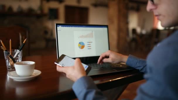 Vista posterior del empresario freelancer en gafas que trabajan en el ordenador portátil con diagramas en la pantalla en la cafetería y comprobar la aplicación de teléfonos inteligentes para obtener información y actualizaciones. Seguimiento y disparo revelador — Vídeos de Stock