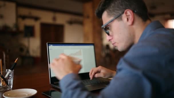 Vue arrièred'un homme d'affaires indépendant travaillant sur un ordinateur portable avec des diagrammes à l'écran dans un café, puis boit une tasse de café. Homme travaillant avec PC dans le coffeeshop. Concept de multitâche d'entreprise . — Video