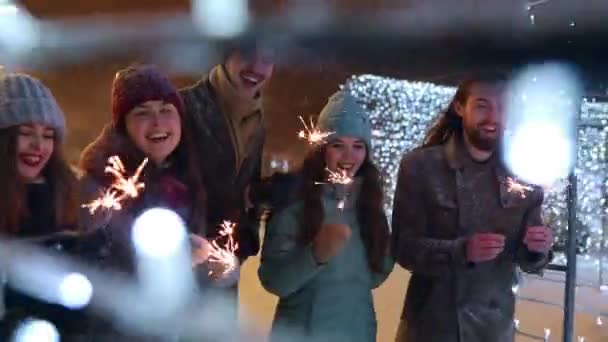 Přátelé se baví procházkou s jiskrami ve slavnostním osvětlovacím tunelu za zasněžené zimní noci. Šťastná skupina lidí visících na vánočním trhu. Garland lampy, kolotoč světla na pozadí. — Stock video