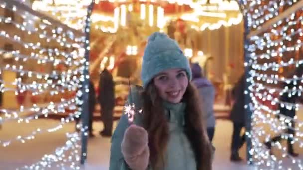 Χαμογελαστή γυναίκα που περπατάει με σπινθήρα σε εορταστική σήραγγα φωτισμού σε χιονισμένη νύχτα. Ένα όμορφο κορίτσι με χειμωνιάτικα ρούχα φλερτάρει στην Χριστουγεννιάτικη αγορά. Λαμπτήρες Garland, φώτα καρουζέλ στο παρασκήνιο. — Αρχείο Βίντεο
