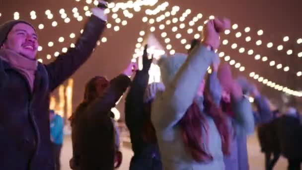 Young people partying at Christmas concert on city square. Friends have fun jumping with raised hands and dancing with arms up at New Year market music stage. Snowfall on winter night celebrations. — Stock Video