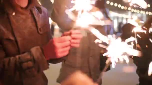 Friends have fun partying with sparklers and doing selfie photo on smartphone at Christmas market. People jump and dance at New Year fairground on winter night. Snowfall, bulb garlands on backdrop. — ストック動画