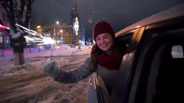 Jovem mulher feliz se inclina para fora da janela do carro e brinca com sparkler na noite de inverno nevado. Menina bonita se diverte na condução de veículos na estrada decorada com luzes de iluminação de Natal e guirlandas bulbo . — Vídeo de Stock