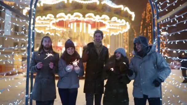 Vrienden hebben plezier wandelen met sterretjes in feestelijke verlichtingstunnel op besneeuwde winternacht. Een gelukkige groep mensen die op de kerstmarkt hangen. slingers, carrousellampjes op achtergrond. — Stockvideo