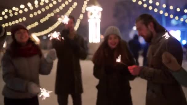 Vrienden hebben plezier feesten met sterretjes en exploderen confetti kanon op de kerstmarkt. Vrolijke mensen springen en dansen op nieuwjaarskermis in de winter besneeuwde nacht, bol bloemenslingers op de achtergrond. — Stockvideo