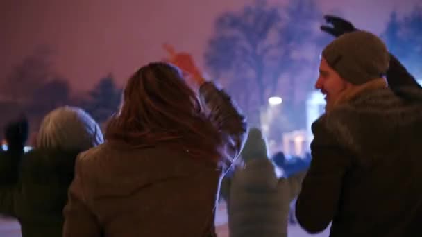 Jóvenes de fiesta en el concierto de Navidad en la plaza de la ciudad. Los amigos se divierten saltando con las manos levantadas y bailando con los brazos en el escenario de música del mercado de Año Nuevo. Nieve en las celebraciones de la noche de invierno . — Vídeo de stock