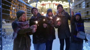 Mutlu arkadaşlar ellerinde maytaplarla yürüyüp neşeli aydınlatma tünelinde karlı kış gecelerinde eğleniyorlar. Noel pazarında takılan insanlar. Çelenk lambaları, arka planda atlıkarınca ışıkları.