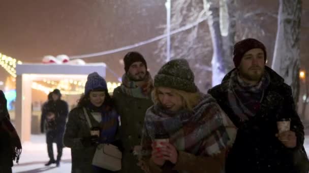 雪の下を歩いている幸せな友人のグループと紙コーヒーカップを手に 雪と遊ぶのを楽しんでください 人々は熱い飲み物で暖まり クリスマスフェアや新年の市場にぶら下がっている — ストック動画