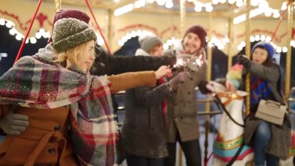在圣诞市场上 微笑的朋友们与骑旋转木马的火星车在一起玩乐 快乐的男男女女们挂在新年的游乐场里 背景上的园林灯 人们在雪夜放松 — 图库视频影像