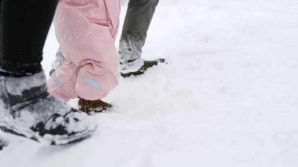 Baby fötter i varm overall gör de första stegen. Barnet lär sig att gå. Vänlig full familj promenader i vinterskogen på semester under snöfall. Far, mor och dotter. Lågvinkelskott. — Stockvideo