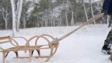 Karlı kış gününde tahta kızak çeken, kalın giysili yakışıklı bir çocuk. Küçük adam kar yağarken parkta kayıyor. Donanma ceketli bir çocuğun ormanda kızakla kaymak için tepeye tırmanırken görüntüsü. Yavaş çekim.