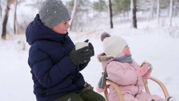 Boy drinks tea and girl eats Christmas gingerbread in jumpsuit after sledding on snowy day. Brother and sister on winter holidays together. Caring father and mother come in composition, hug, kiss kids — Stock Video