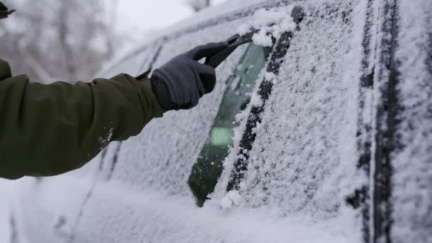 Čistí boční okna auta od sněhu s škrabkou na led před cestou. Člověk odstraňuje led z oken auta. Mužská ruka čistí auto se speciálním nářadím na zasněžený zimní den. Zpomalený pohyb 120 fps. — Stock video