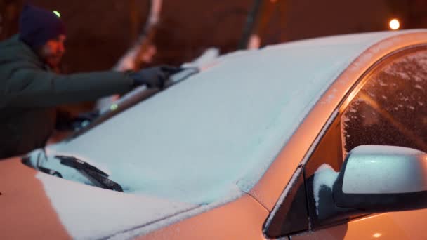 Reiniging van de zijruiten van de auto van sneeuw met ijsschraper voor de reis. De man haalt ijs uit autoruiten. man reinigt auto op besneeuwde winternacht. — Stockvideo