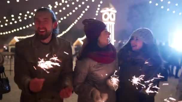 Cheerful friends have fun partying with sparklers in hands at Christmas market. People jump and dance at New Year fairground on winter night with snowfall, bulb garlands on backdrop. Birthday party. — ストック動画