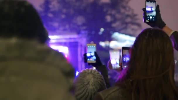 Crowd on Christmas concert recording video and taking photos on smartphone camera in raised hands near stage. Young people partying at New Year on city square musical show. Snowy winter night. — ストック動画