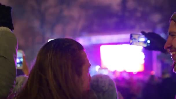 クリスマスコンサートのレコーディングビデオに群衆やステージ近くの調達手でスマートフォンのカメラで自撮り写真を撮る。市の広場のミュージカルショーで新年を祝う若い人たち。冬の雪の夜. — ストック動画