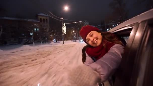 若い幸せな女性は車の窓から傾き、雪の冬の夜に輝くと遊ぶ。かわいい女の子はクリスマスイルミネーションと電球のガーランドで飾られた道路上で車を運転するのが楽しいです. — ストック動画