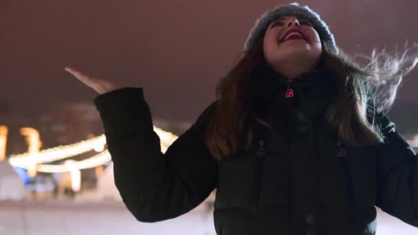 Mujer joven disfrutando de la caída de nieve al aire libre en la noche de invierno en el mercado de Navidad. Chica feliz abrir los brazos bajo la nieve en el parque y coger los copos de nieve con las manos, la boca. Mujer estirar los brazos al cielo . — Vídeos de Stock