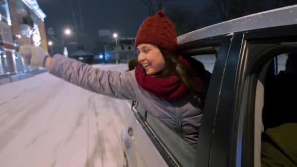 Junge glückliche Frau lehnt sich aus dem Autofenster und spielt in einer verschneiten Winternacht mit Wunderkerzen. hübsches Mädchen hat Spaß beim Autofahren auf der Straße mit Weihnachtsbeleuchtung und Glühbirnengirlanden dekoriert. — Stockvideo
