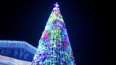 Karlı kış gecelerinde şehir meydanında parlayan parlak çelenklerle süslenmiş büyük bir Noel ağacı. Dışarıdaki dekorasyonlarla dolu yeni yıl ağacının etrafında kameralar uçuşuyor. Işık Gösterisi.