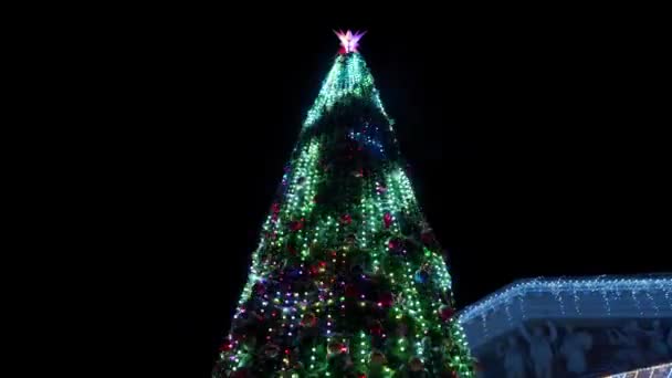 Grande árvore de Natal decorada com guirlandas luminosas piscando na praça da cidade na noite de inverno nevado. Câmera voa ao redor brilhante árvore de Ano Novo cheio de decorações ao ar livre na rua. Mostra de luz . — Vídeo de Stock