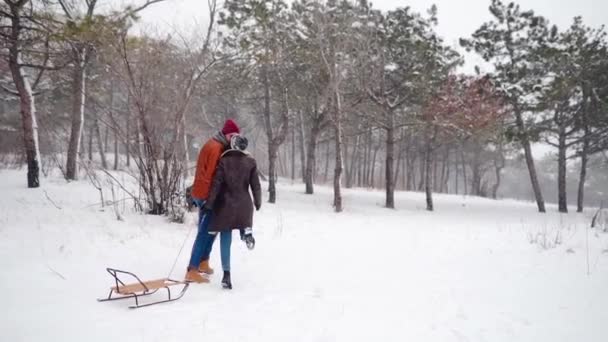 Прекрасна пара ходить, тягнучи санки в сніжний зимовий день. Чоловік з дівчиною сідає на сніг. Жінка їде кататися на свіжому повітрі з хлопцем. Людям подобаються різдвяні канікули. Повільний рух . — стокове відео