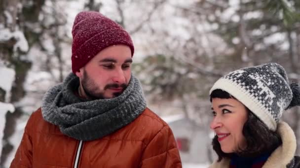 Unga kärleksfulla par går i en park på snöfall. Mannen och hans flickvän håller hand och njuter av snö på vinterdagen. Hipster kvinna promenera i snöig skog leende med pojkvän i snygga kläder. — Stockvideo