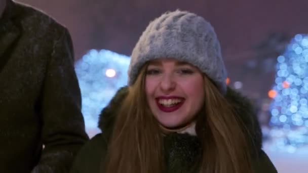 Vrienden wandelen onder sneeuw op kerstmarkt op besneeuwde winternacht op kermis. Een groep gelukkige mensen die rondhangen, plezier hebben en lachen. Sneeuwval en glanzende bloemenslingers op de achtergrond — Stockvideo