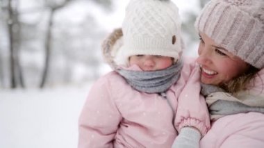 Anne, Noel tatilinde eşofmanlı kızını açık havada karlı bir günde taşıyor. Kışın kar altında pembe tulumlarda bebekle yürüyen bir kadın. Sıcak giysili insanlar. Yavaş çekim.