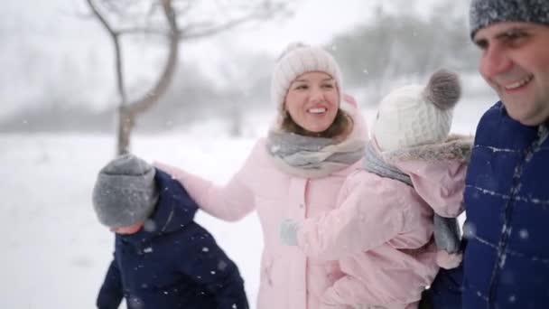 Friendly full family walking through winter forest on vacation under snowfall. Father, mother, son and daughter rest on Christmas holidays outdoors on snowy day. People in warm clothes. Slow motion. — Stock Video