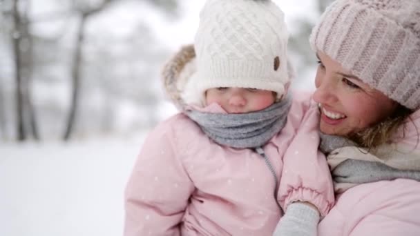 Mor bär dotter i overall på jullov utomhus på snöig dag. Kvinna promenader med barn i rosa sparkar i vinterskogen under snöfall. Folk i varma kläder. Långsamma rörelser. — Stockvideo