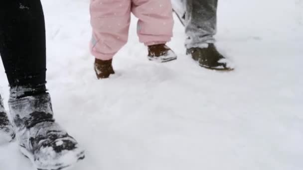 Pies de bebé en mono caliente dando los primeros pasos. El niño aprende a caminar. Amistosa familia completa caminando en el bosque de invierno de vacaciones bajo las nevadas. Padre, madre e hija. Disparo de ángulo bajo . — Vídeo de stock