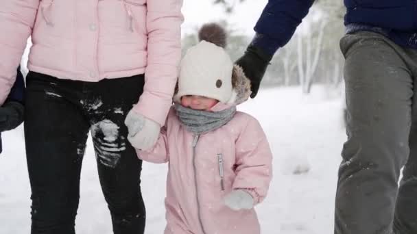 Baby feet in warm jumpsuit doing first steps. Child learns to walk. Friendly full family walking in winter forest on vacation under snowfall. Father, mother, son and daughter. Low angle shot. — Stock Video