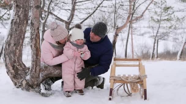 Маленькая девочка в розовом зимнем комбинезоне ест рождественские пряники после катания на санках в снежный день. Прекрасная семья проводит зимние каникулы вместе. Заботливый отец и мать объятия, поцелуй очаровательный ребенок . — стоковое видео