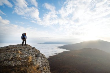 Sırt çantalı genç bir turist dağın tepesinde duruyor ve bulutların üzerinde güzel sarı sonbahar günbatımını seyrediyor.