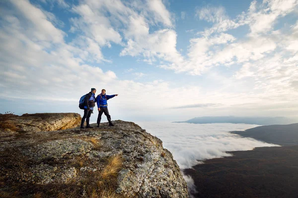 Two climbers standing on top of summit above clouds in the mountains. Hiker man pointing with his hand discussing route. Plan, vision and mission concept.